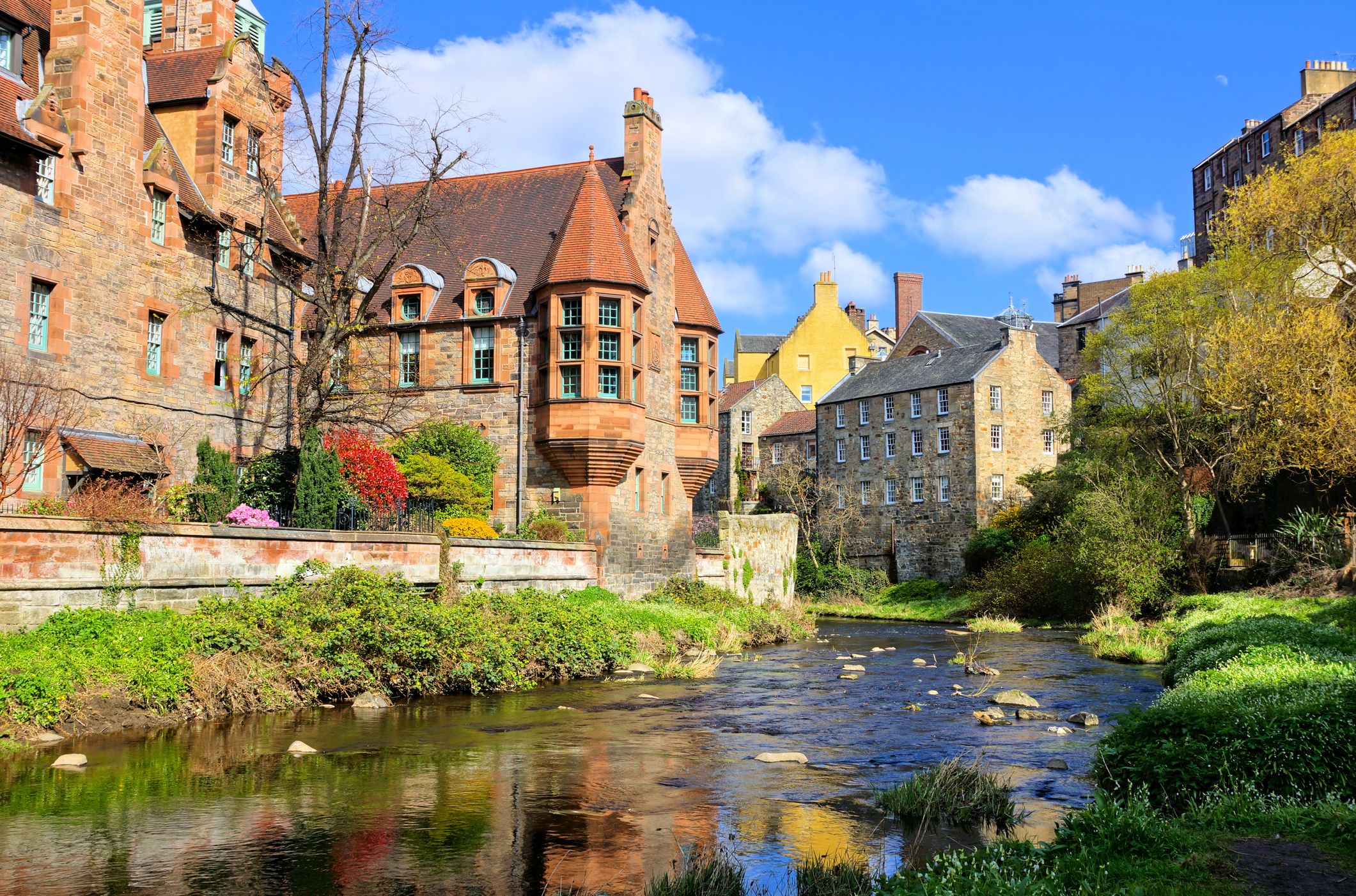 A scenic view of Dean Village in Edinburgh, Scotland, featuring historic stone buildings, lush greenery, and a gently flowing river under a bright blue sky.