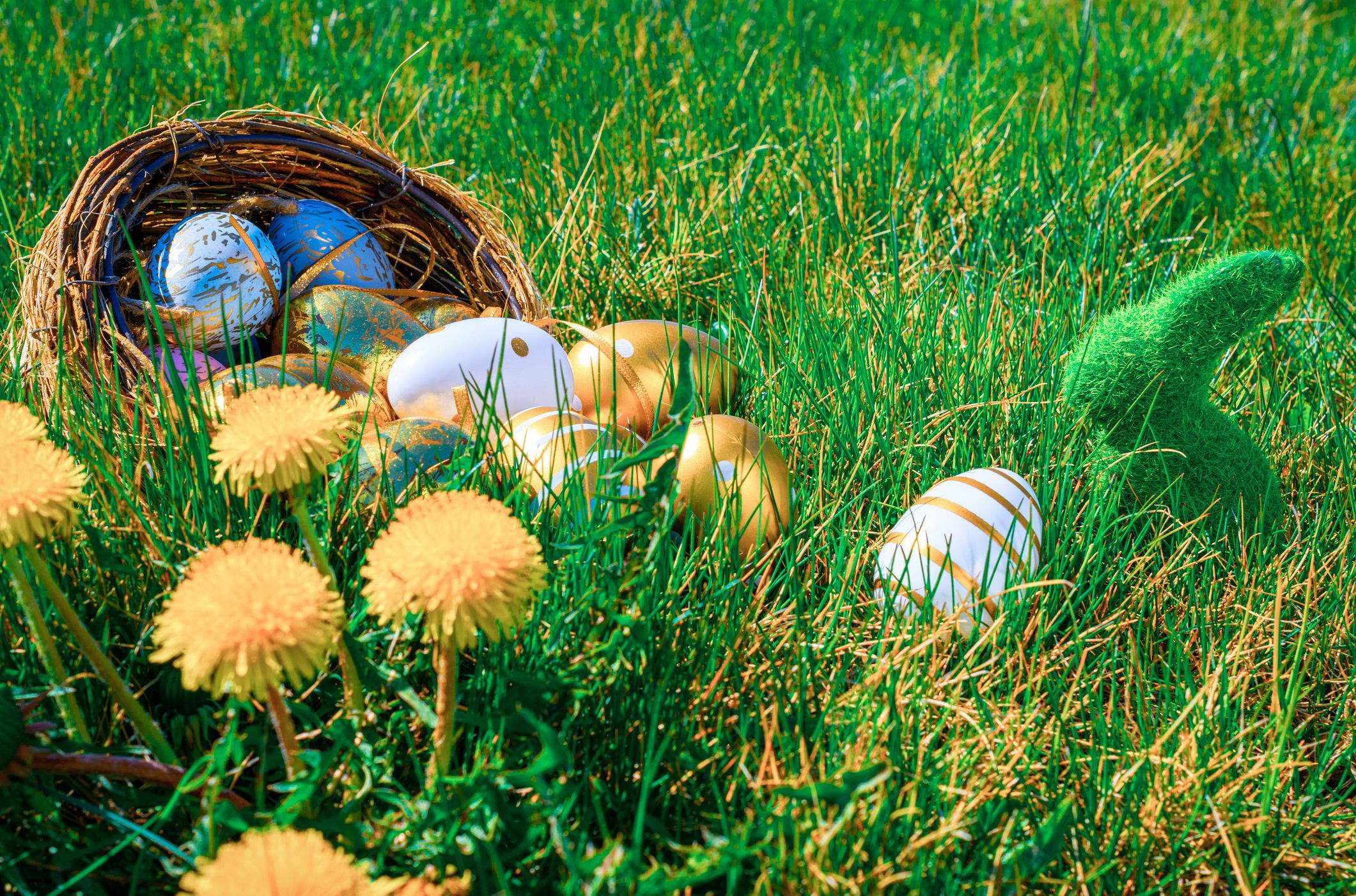 A wicker basket spills golden and decorated Easter eggs onto green grass, surrounded by dandelions, with a fuzzy green bunny figurine nearby.