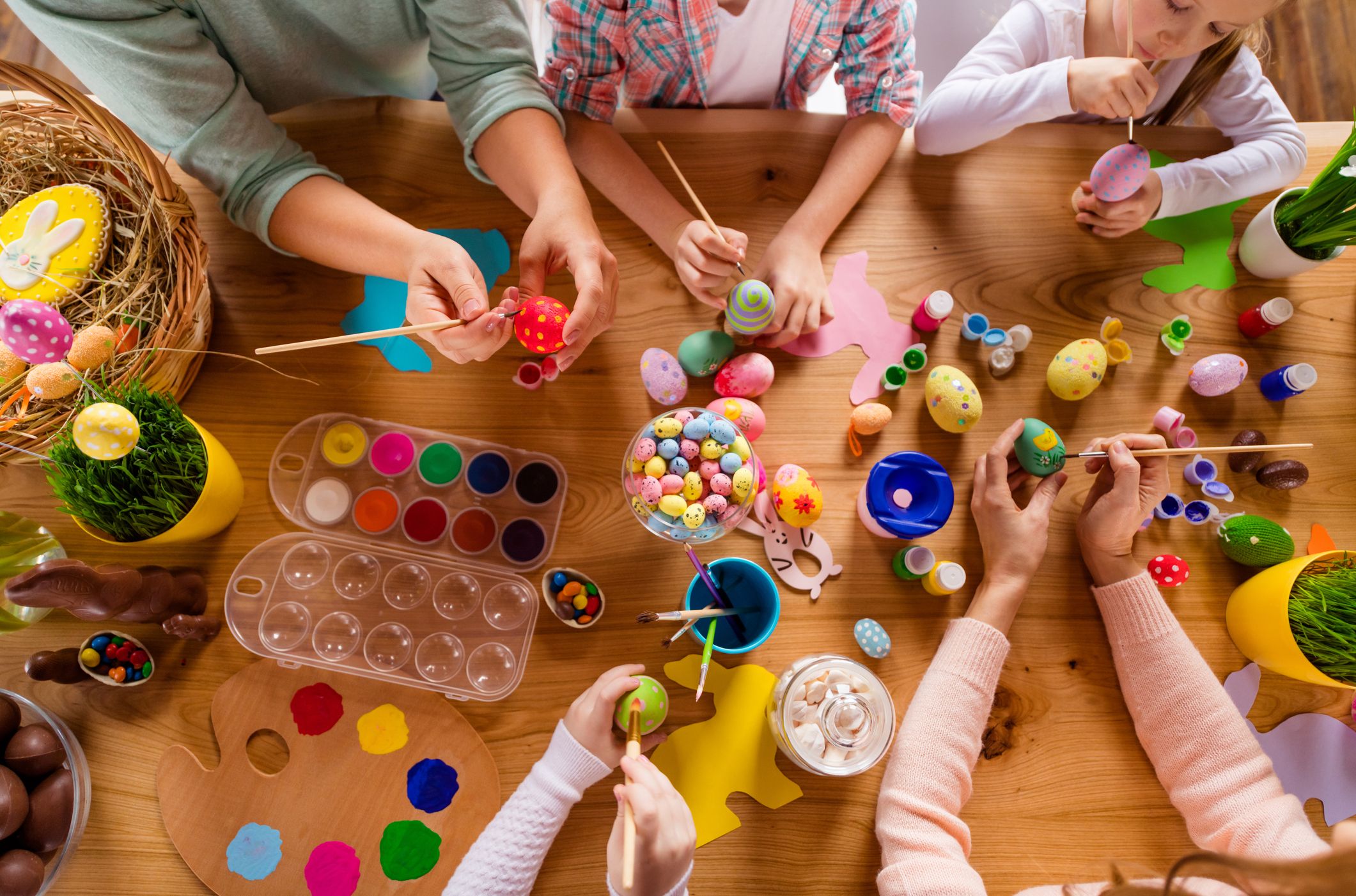 Children and adults gather around a wooden table, painting colorful Easter eggs with brushes and dyes, surrounded by art supplies, candies, and festive decorations.