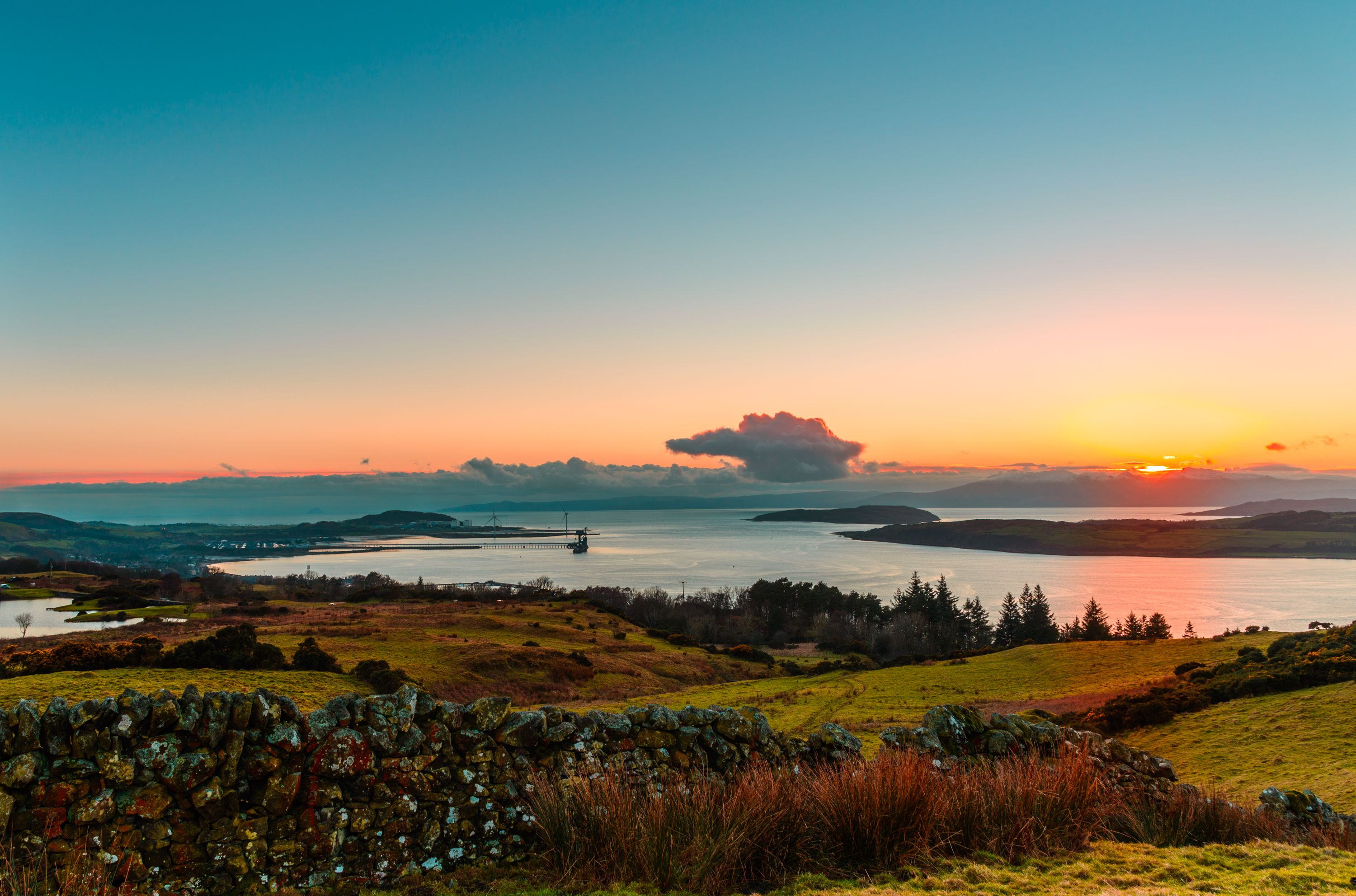 A breathtaking sunset over the Scottish countryside, with rolling green hills, a stone wall in the foreground, and calm waters reflecting the warm hues of the setting sun