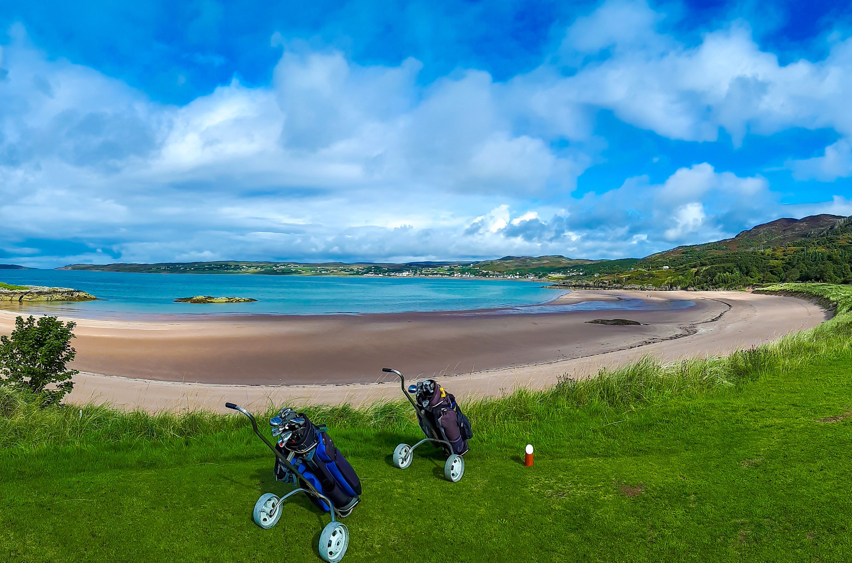 A scenic golf course overlooking a sandy beach and blue waters, with two golf trolleys in the foreground. Lush green hills and a bright sky with clouds complete the view.