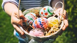 A child holds a wicker basket filled with colorful, hand-painted Easter eggs resting on straw, with a blurred green lawn in the background.