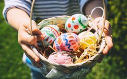 A child holds a wicker basket filled with colorful, hand-painted Easter eggs resting on straw, with a blurred green lawn in the background.