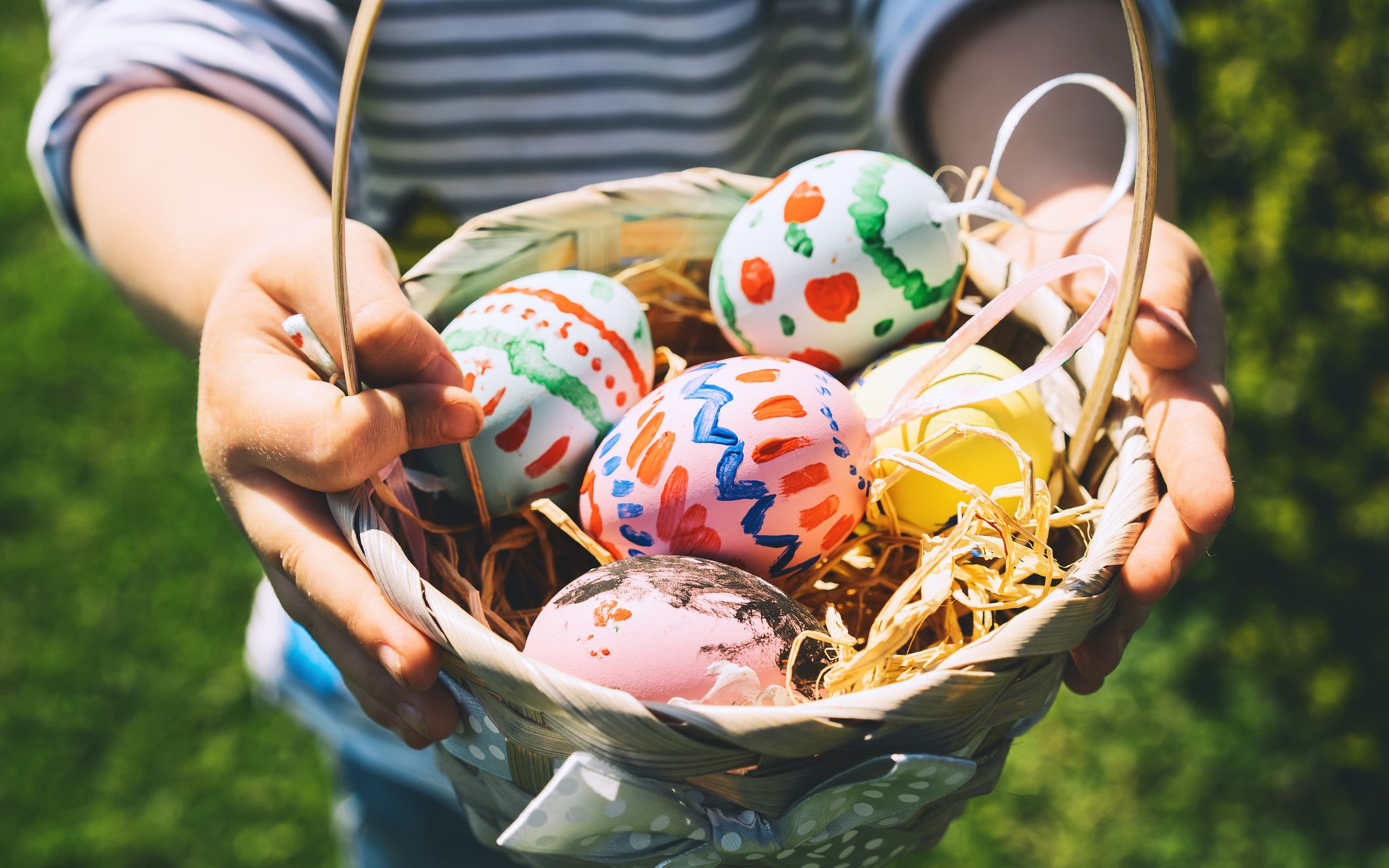 A child holds a wicker basket filled with colorful, hand-painted Easter eggs resting on straw, with a blurred green lawn in the background.