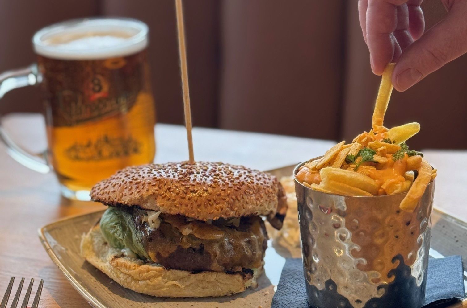 Someone pinching a chip from a plate with a burger and chips on it, with a beer in the background.