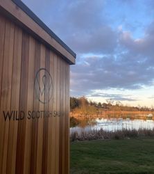 A photograph of the side of the sauna at the resort with the Wild Scottish Sauna hand painted logo on the side, looking out to peaceful pools.