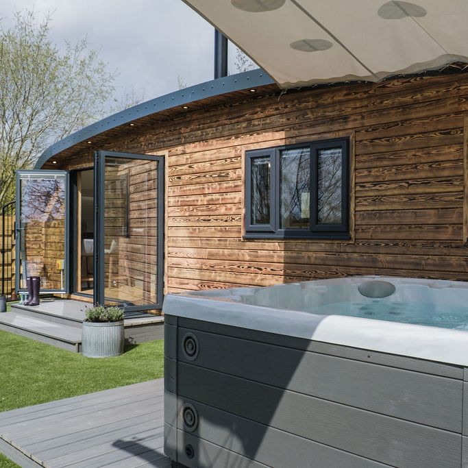 Looking towards an Escape Pod with a fired wood exterior, with a hot tub bubbling beside it.