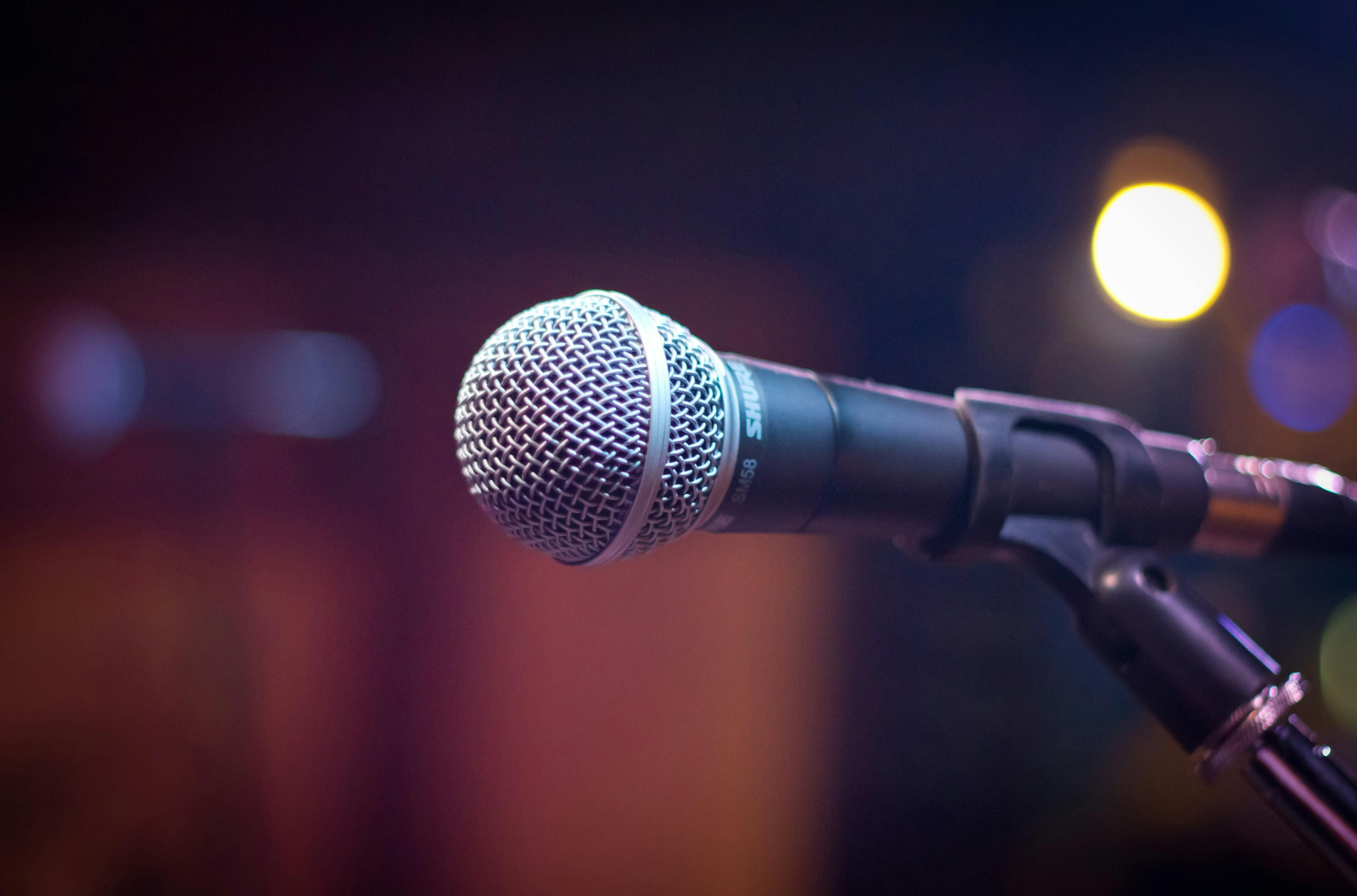 Close-up of a microphone on a stand with blurred stage lights in the background, suggesting a live performance or public speaking setup.
