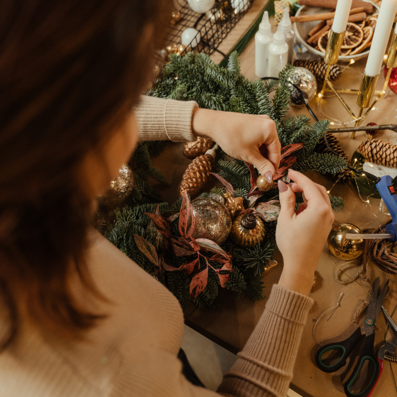 A woman crafting a wreath with a selection of materials from ribbons to rustic pine cones.