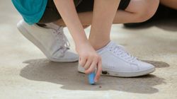 Child crouched on pavement drawing with blue chalk, wearing white sneakers and a turquoise shirt; faint chalk flower outlines visible on the ground.