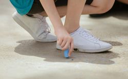 Child crouched on pavement drawing with blue chalk, wearing white sneakers and a turquoise shirt; faint chalk flower outlines visible on the ground.