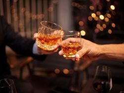 Two people clink glasses of whiskey in a celebratory toast, with warm lighting and festive bokeh in the background.