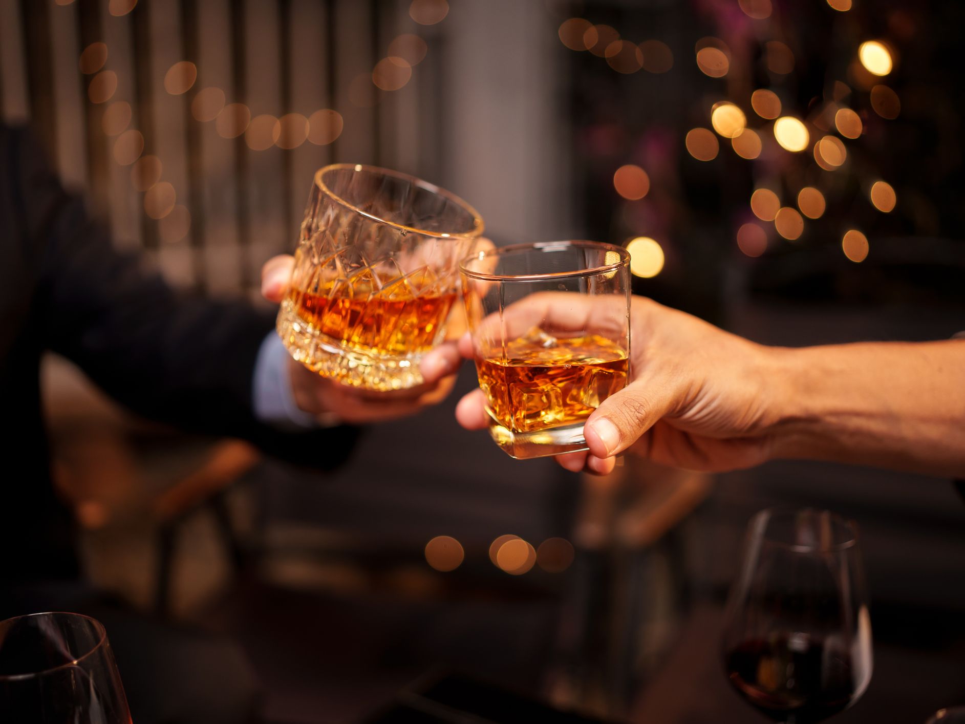 Two people clink glasses of whiskey in a celebratory toast, with warm lighting and festive bokeh in the background.