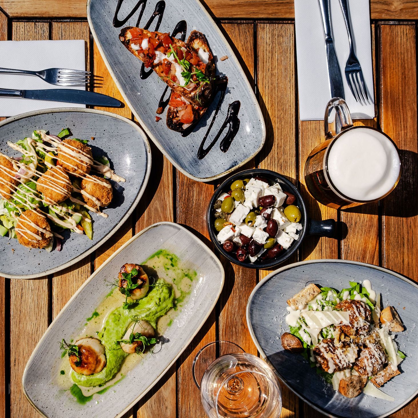 Assorted dishes including salad, bruschetta, scallops, fried appetizers, and olives with cheese on a wooden outdoor table with drinks