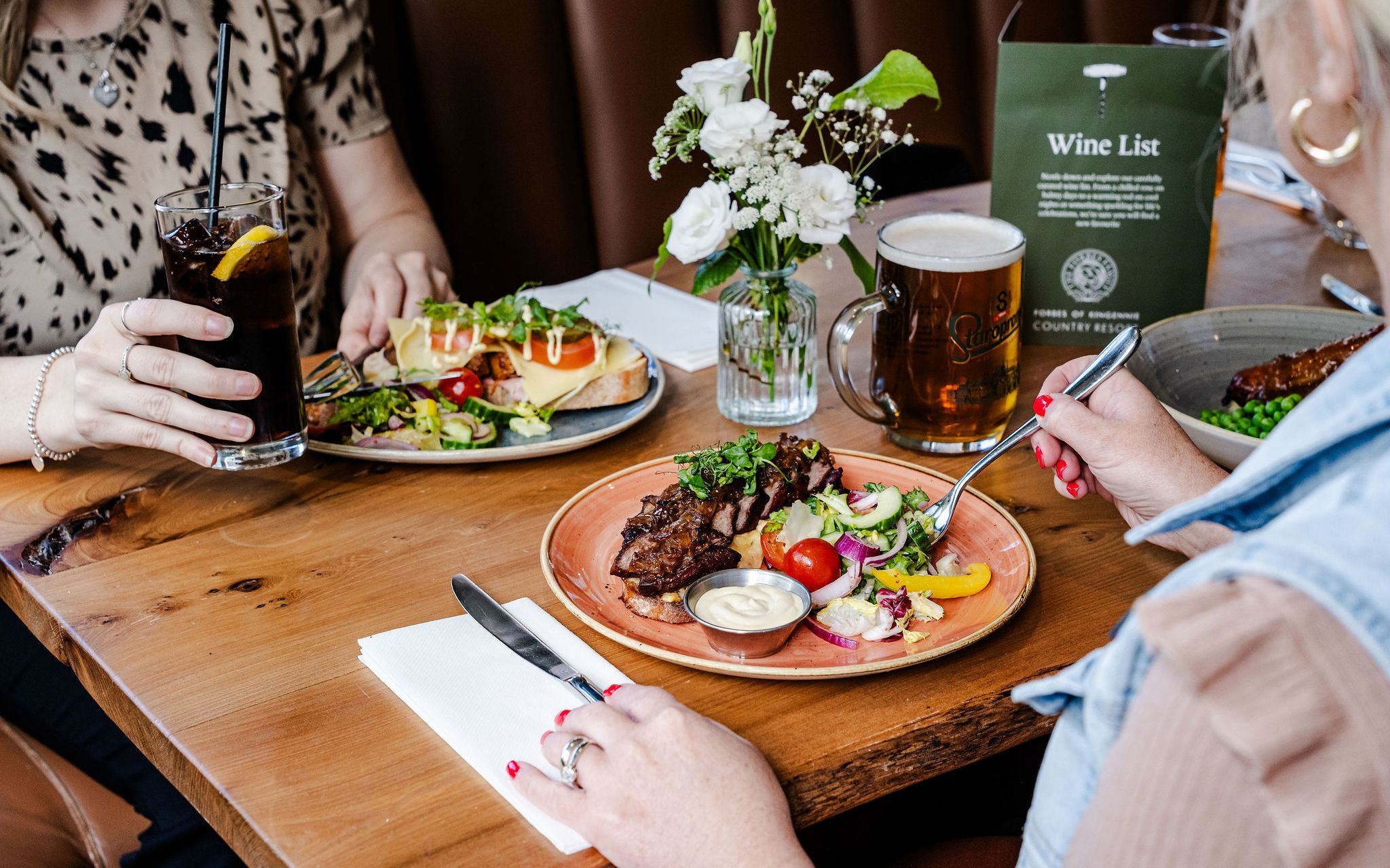People dining at a restaurant table with plates of food, drinks, and a flower vase.