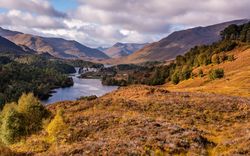 Scenic view of a lake surrounded by autumnal hills and forested mountains under a partly cloudy sky in the Scottish Highlands.