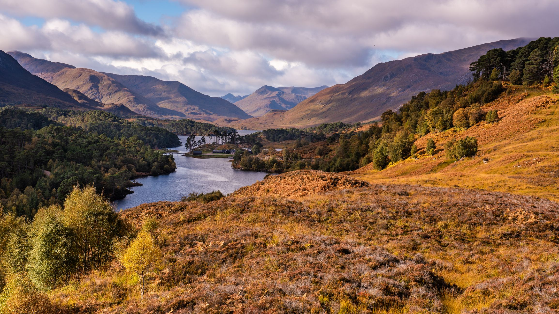 Scenic view of a lake surrounded by autumnal hills and forested mountains under a partly cloudy sky in the Scottish Highlands.