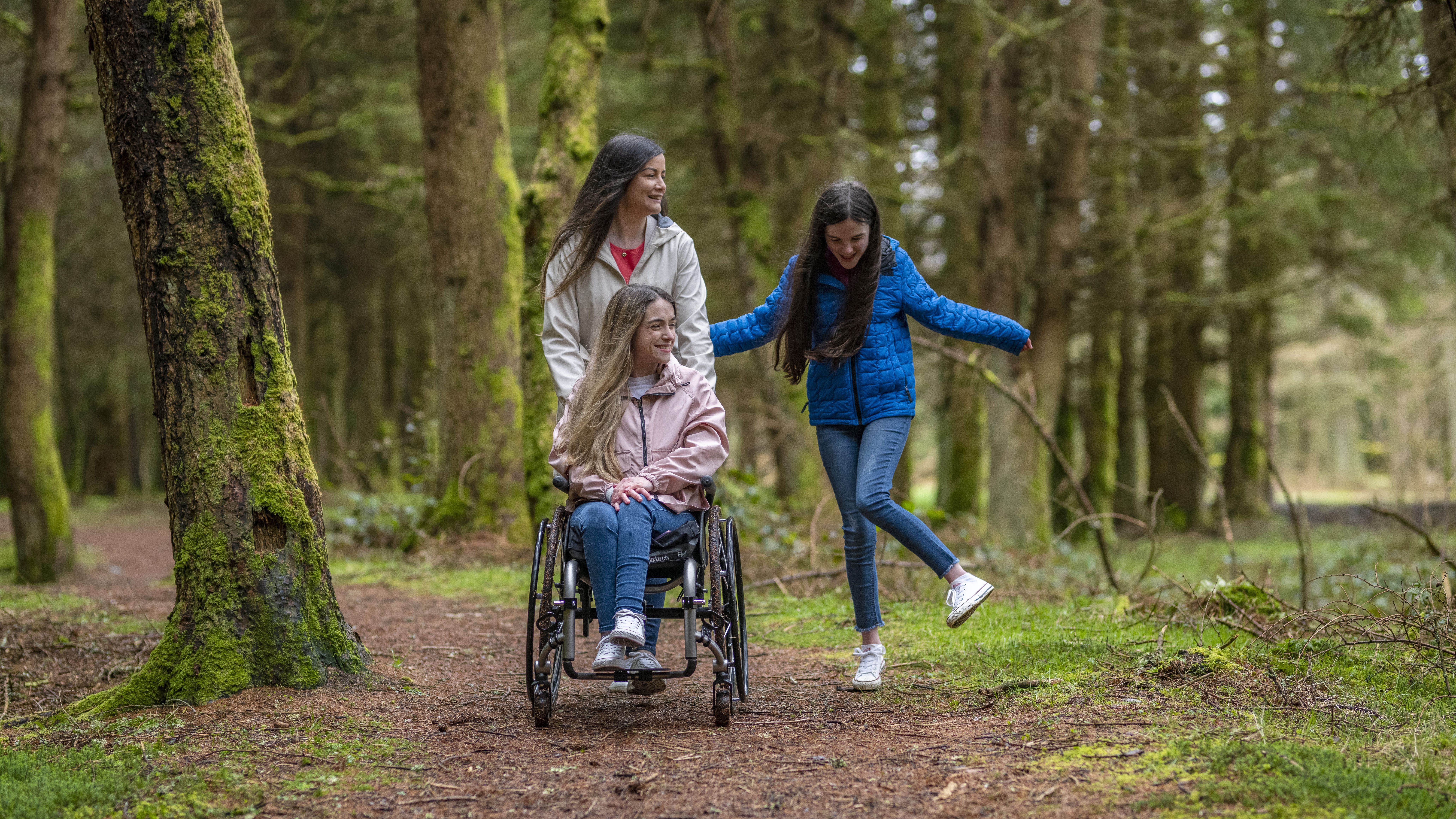 Three women enjoying a walk in a forest, one of whom is in a wheelchair.