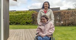 A woman pushing another woman in a wheelchair outside on a wooden deck.