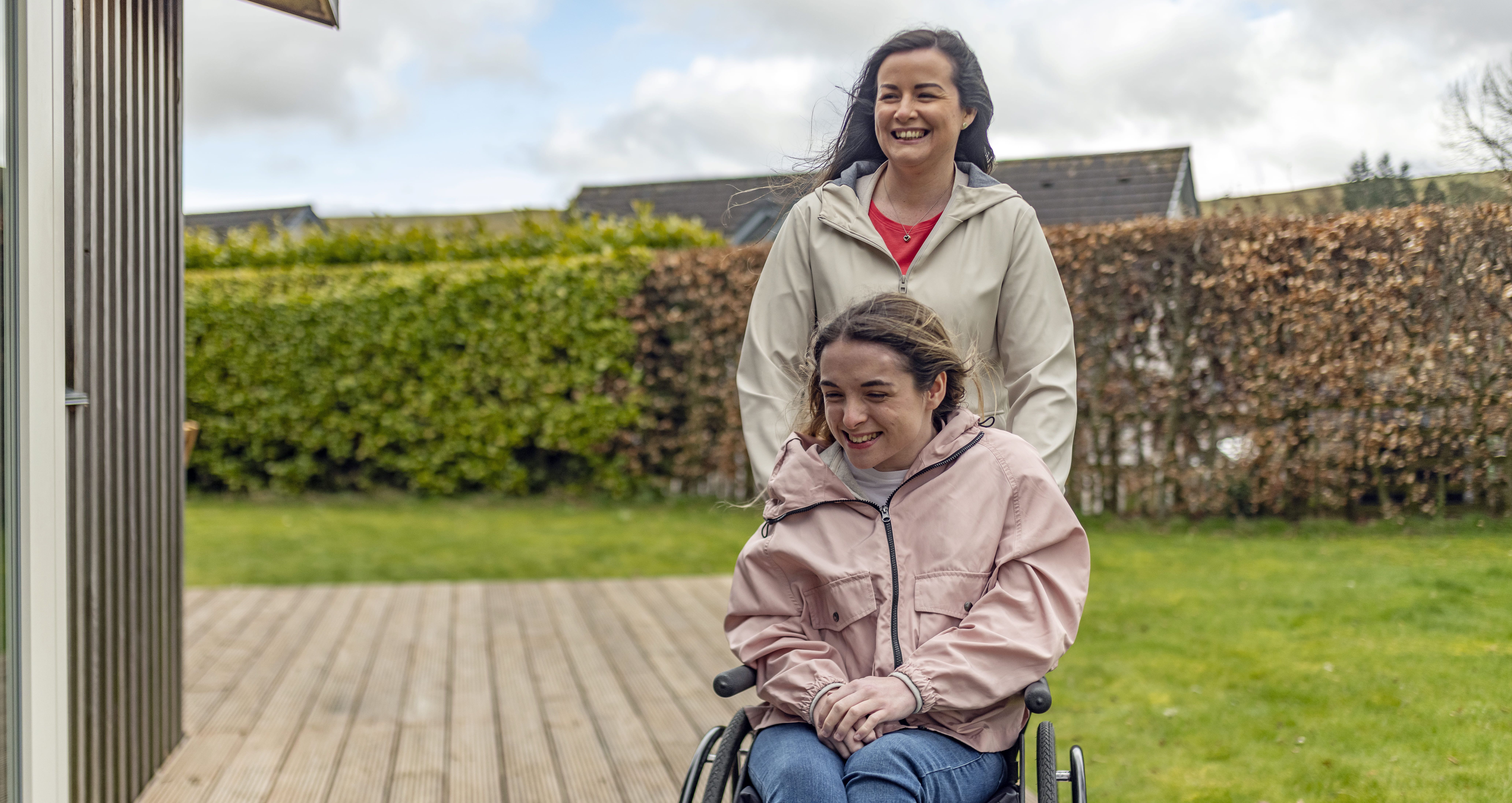 A woman pushing another woman in a wheelchair outside on a wooden deck.