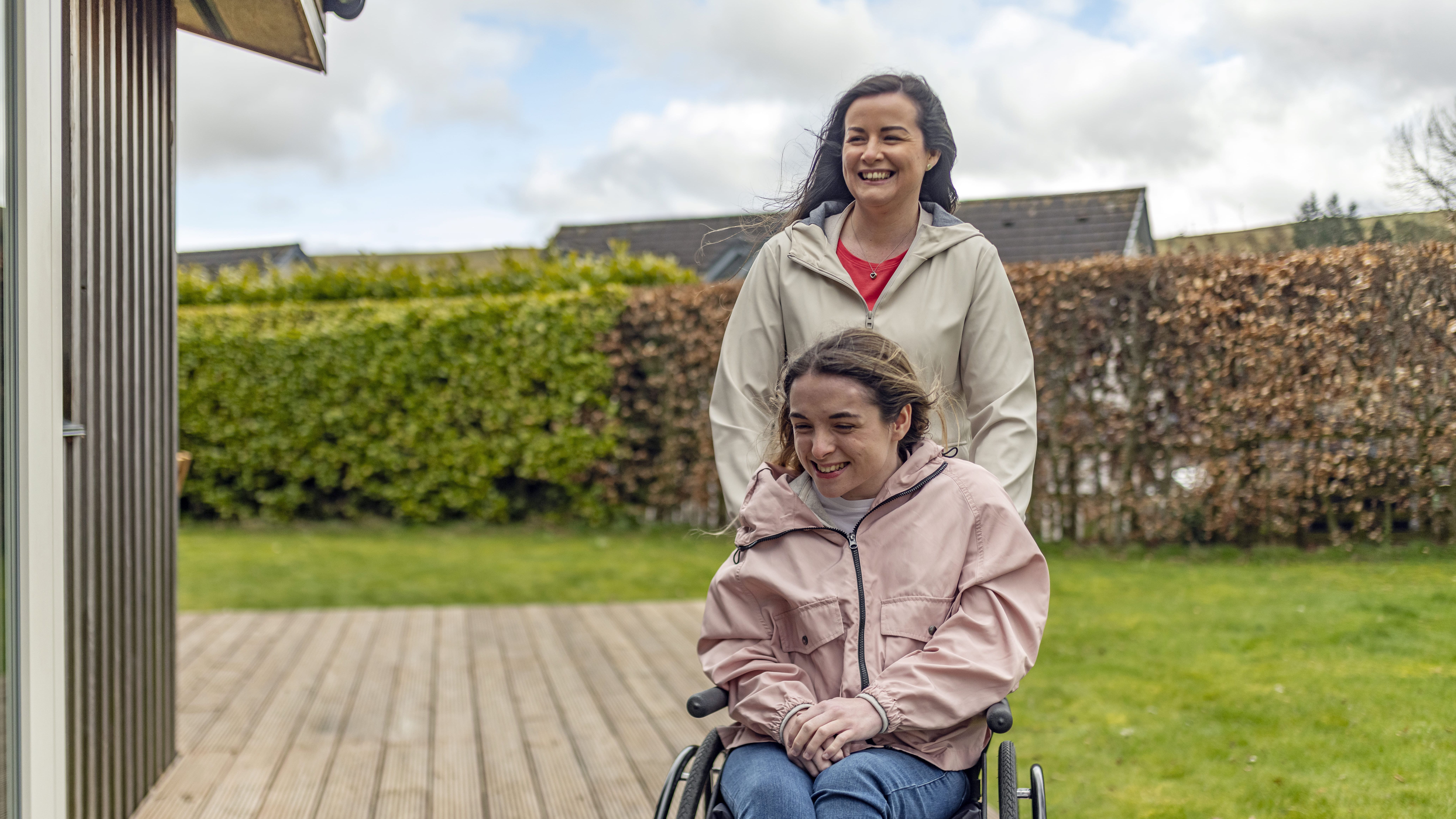 A woman pushing another woman in a wheelchair outside on a wooden deck.