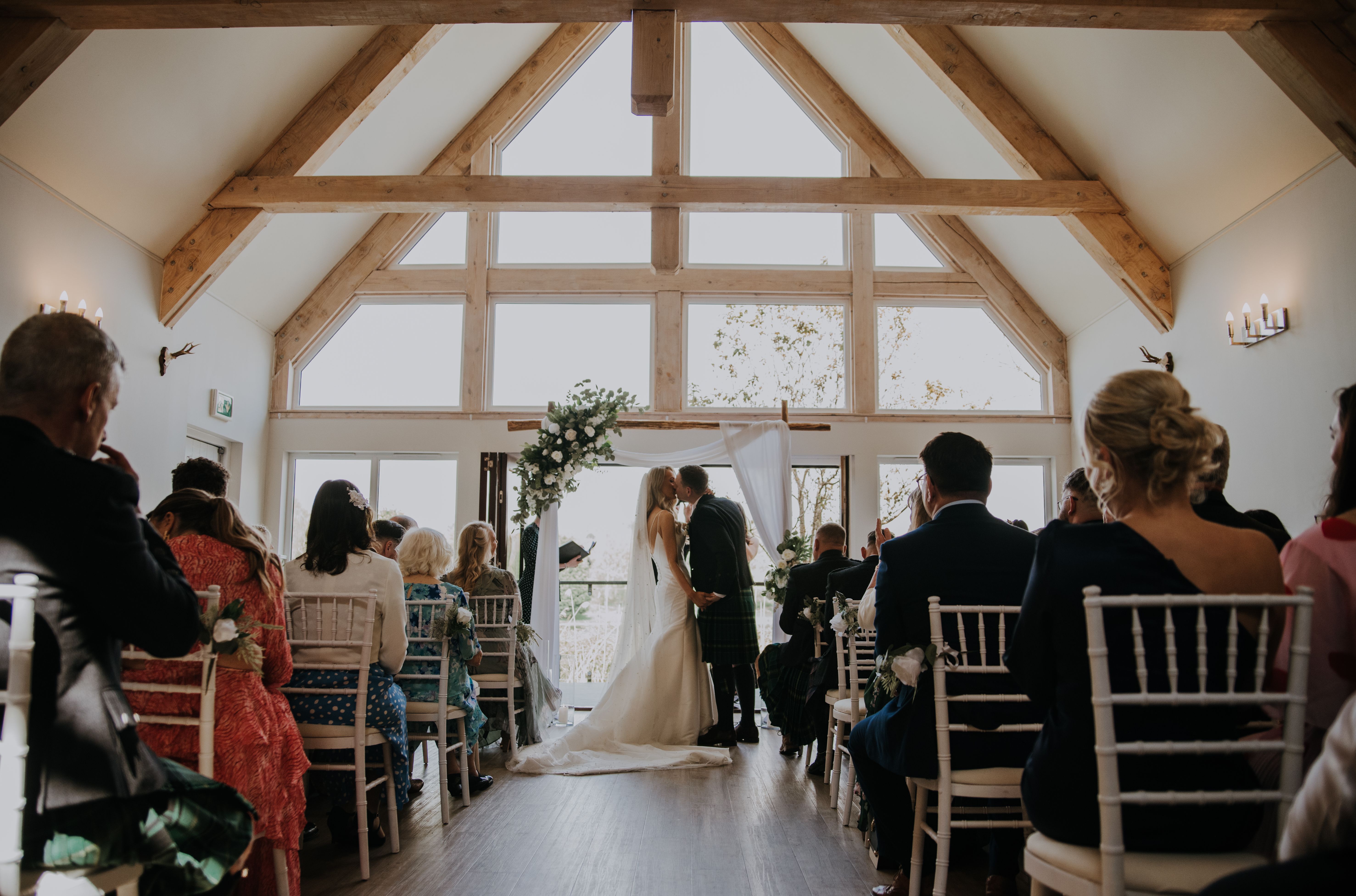 Bride and groom kiss during their wedding ceremony in a bright, wooden-beamed hall filled with seated guests.
