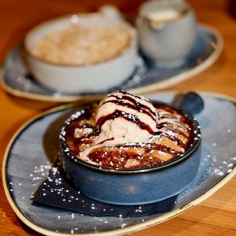 A warm dessert topped with vanilla ice cream and chocolate sauce, served in a small bowl with another dessert in the background.