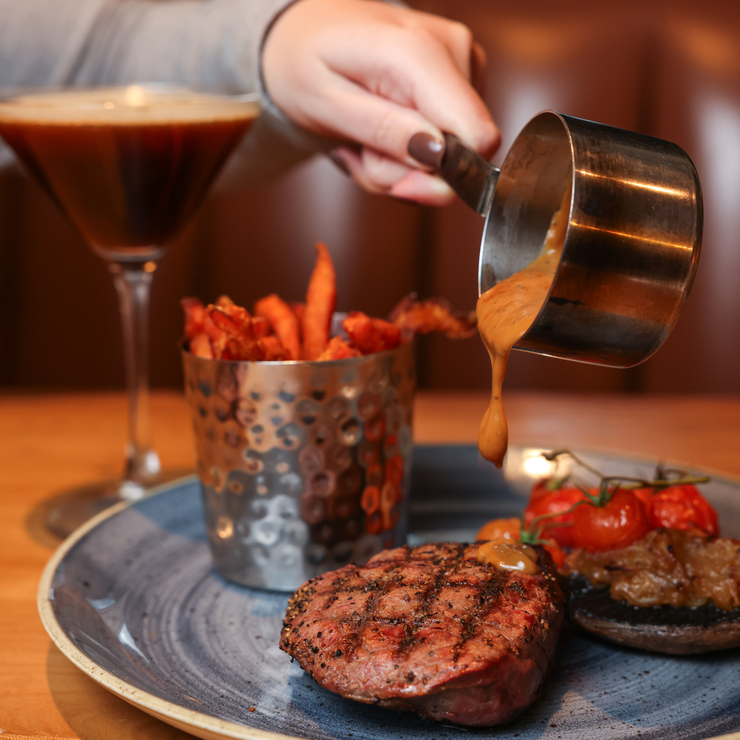 Close-up of a steak dinner being served with sauces, sweet potato fries, grilled tomatoes, and a drink.