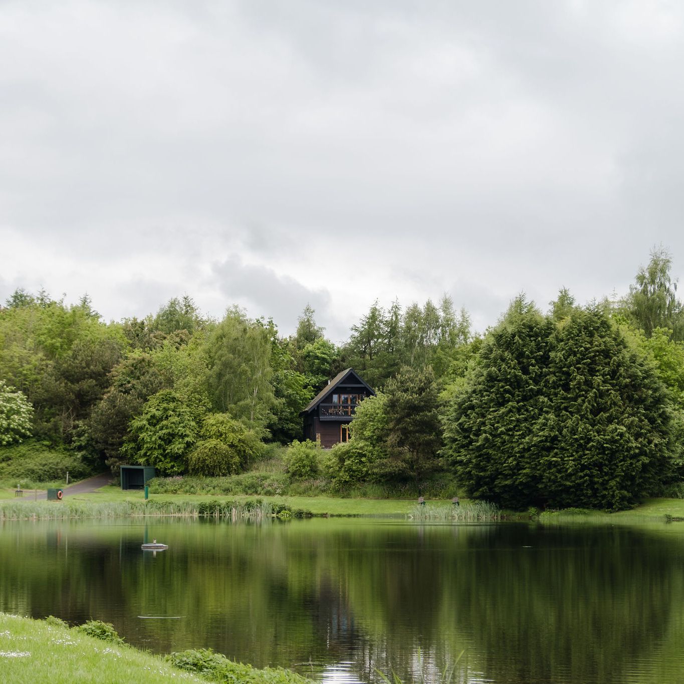 A wooden cabin surrounded by dense green trees, located on a small hill by the edge of a calm lake with reflections of the trees and cabin in the water under a cloudy sky.