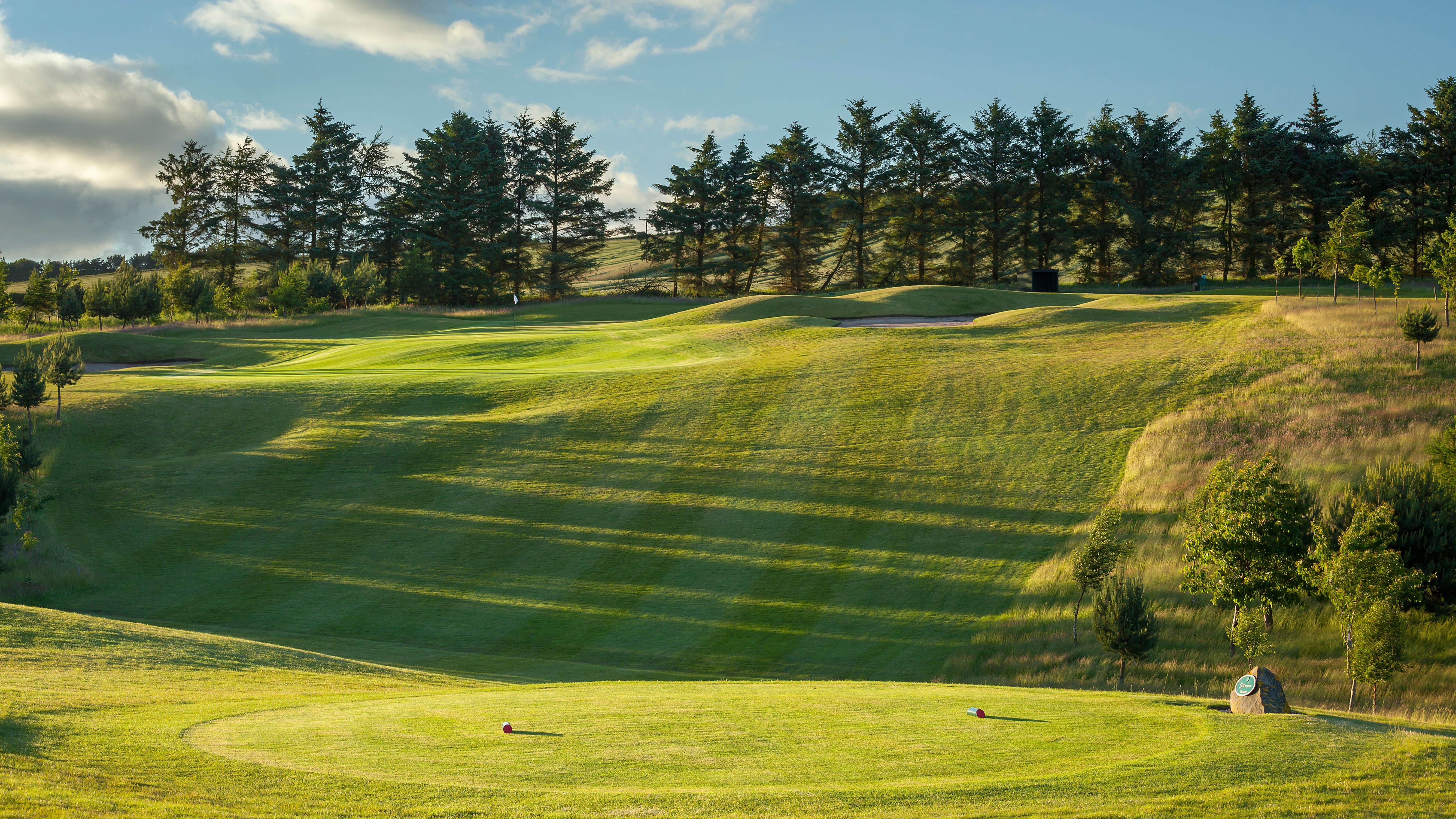 Golf course with green fairways, tee markers, and trees under a blue sky with clouds.
