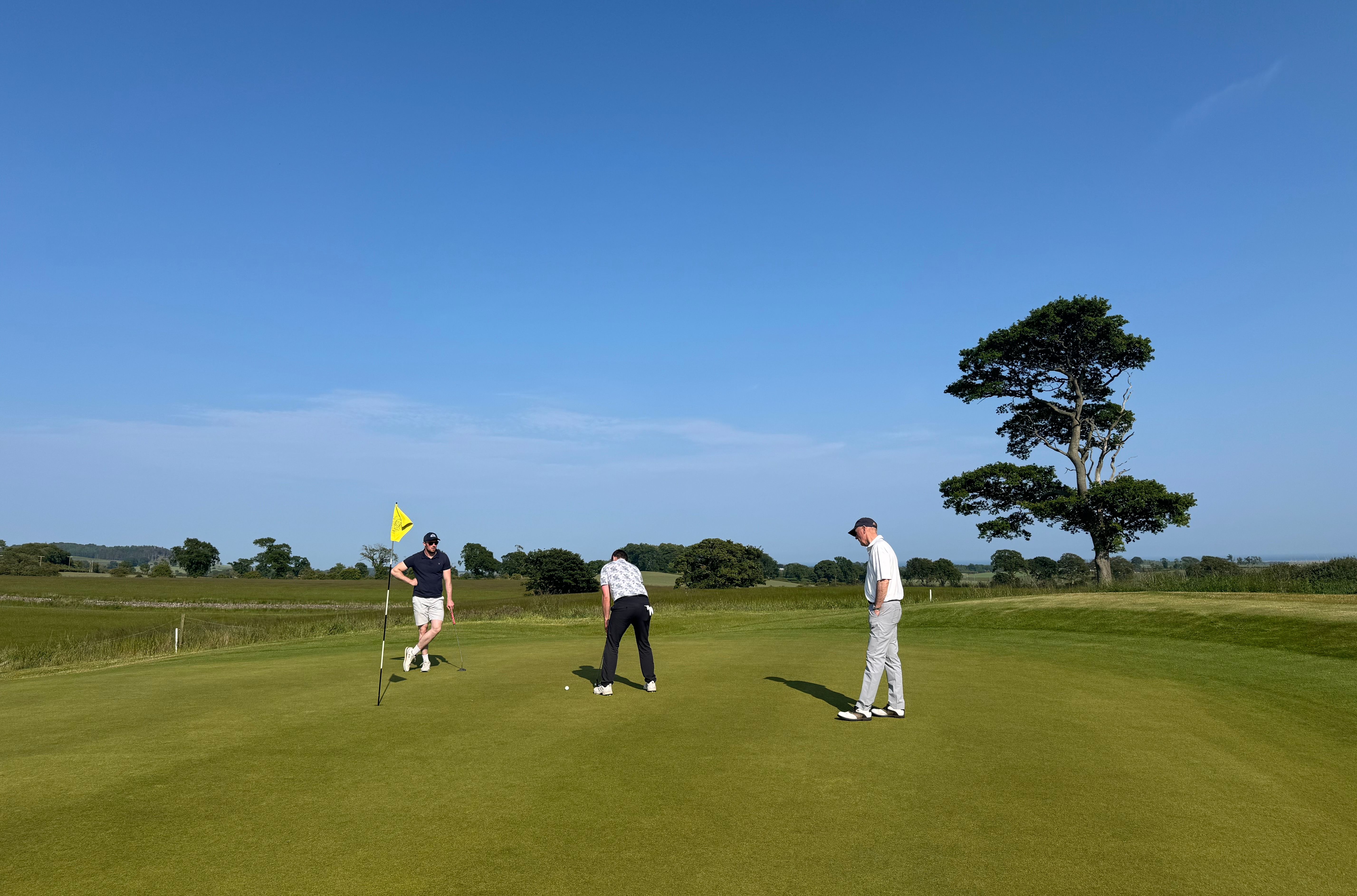 Three people playing golf on a green golf course under a clear blue sky.