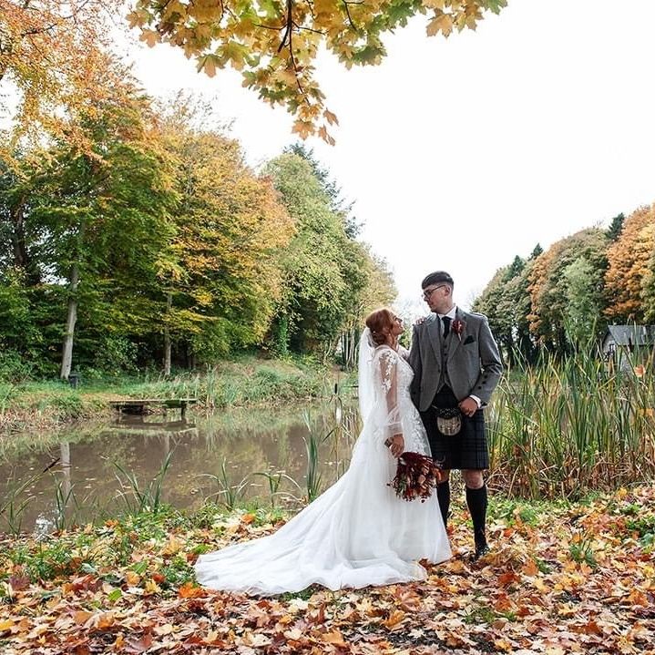 Bride and groom standing by a pond surrounded by autumn trees and fallen leaves