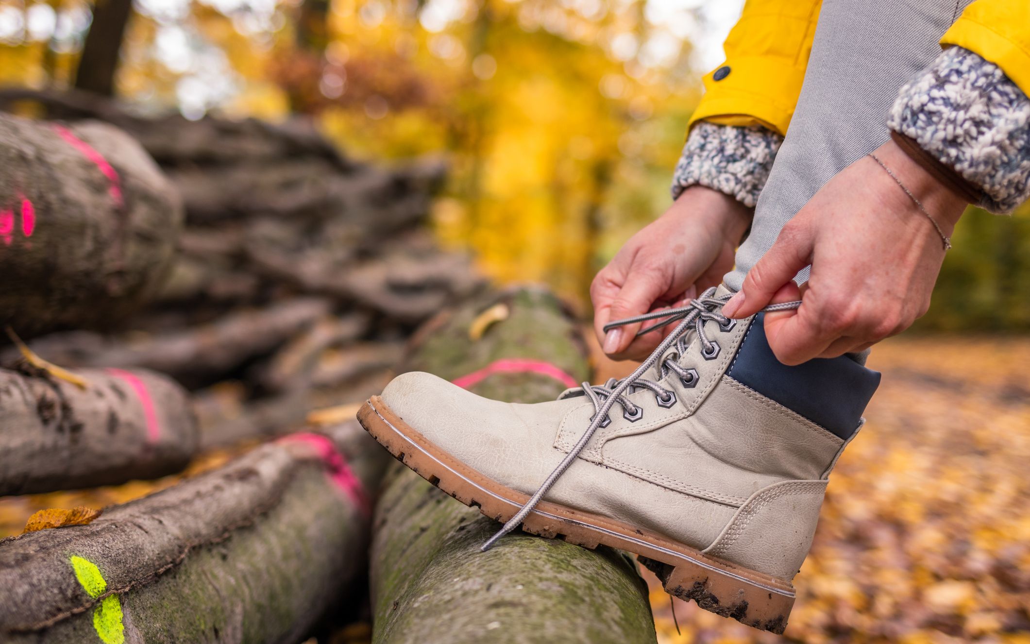 Close-up of a person in a yellow jacket tying a light-colored hiking boot on stacked logs in an autumn forest.