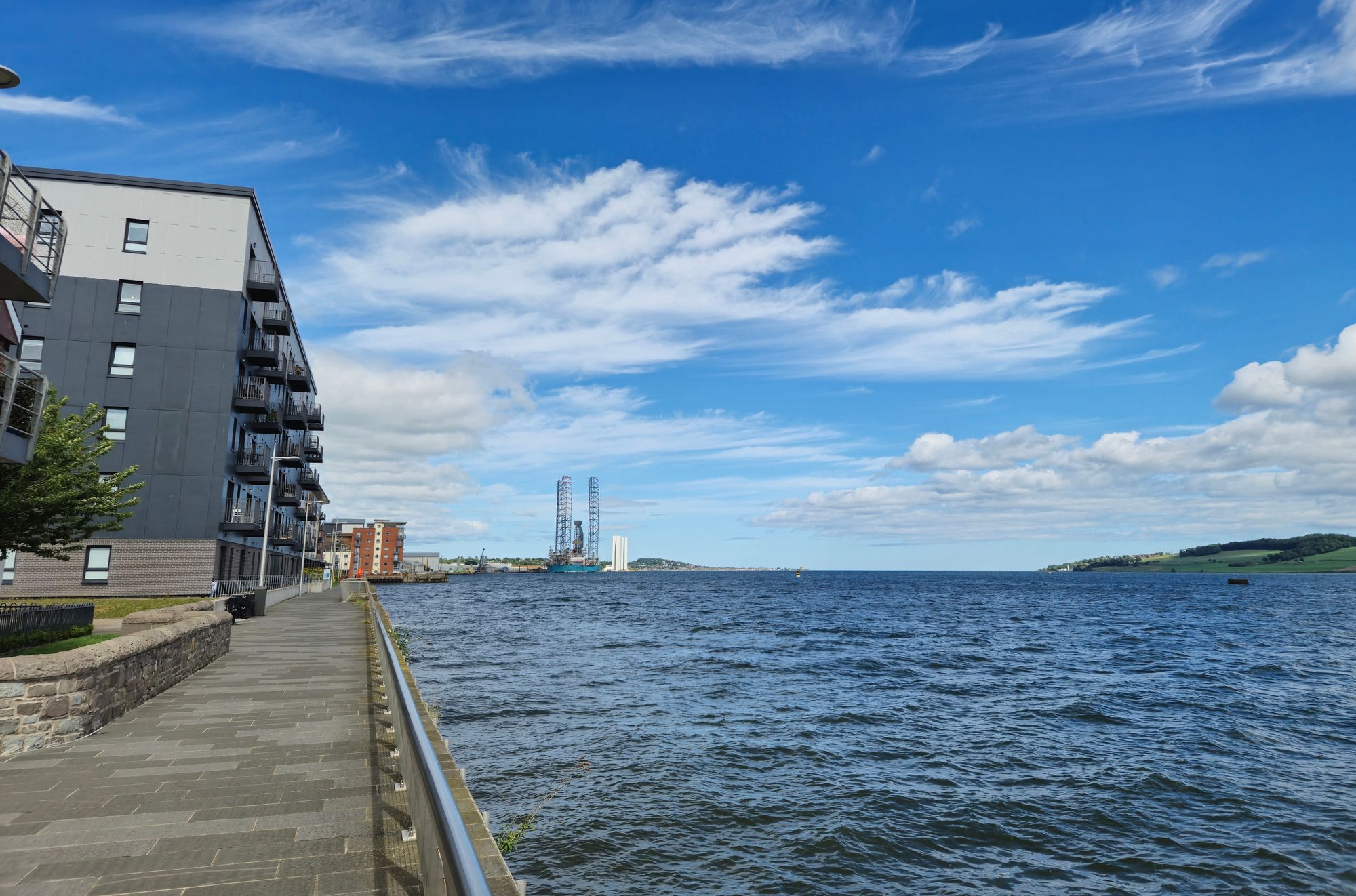 Waterside promenade beside the River Tay in Dundee, with apartment buildings on the left and a wide blue sky over the river.