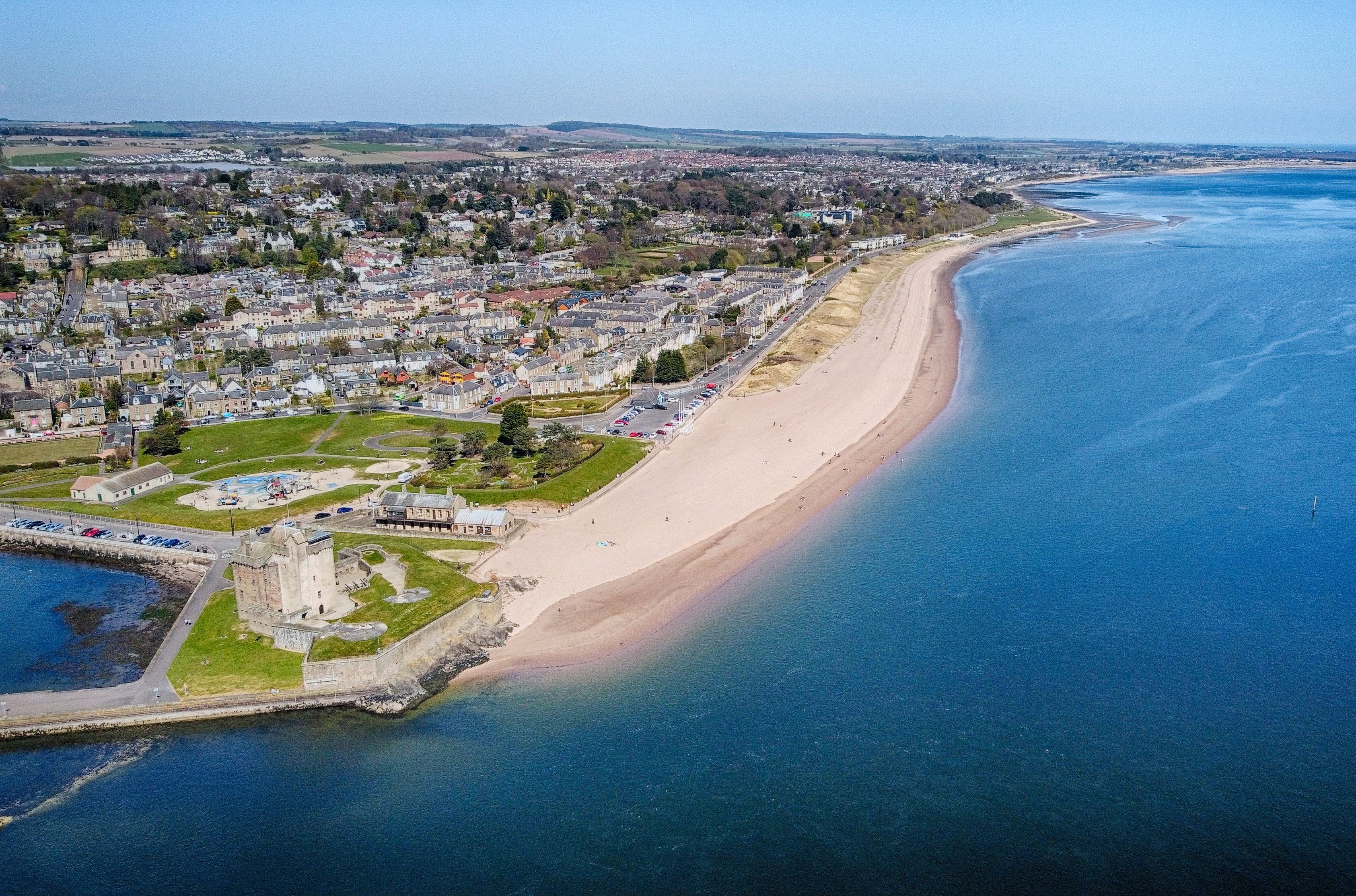 Aerial view of Broughty Ferry in Dundee, showing the beachfront, calm blue water, and Broughty Castle at the edge of the shore.