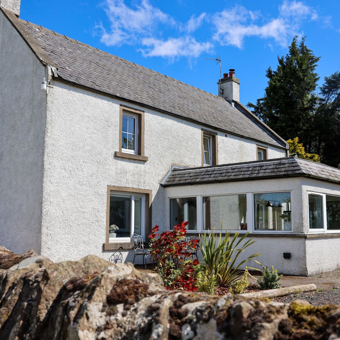 White two-story house with a grey tiled roof, a conservatory, and a stone garden wall in front.