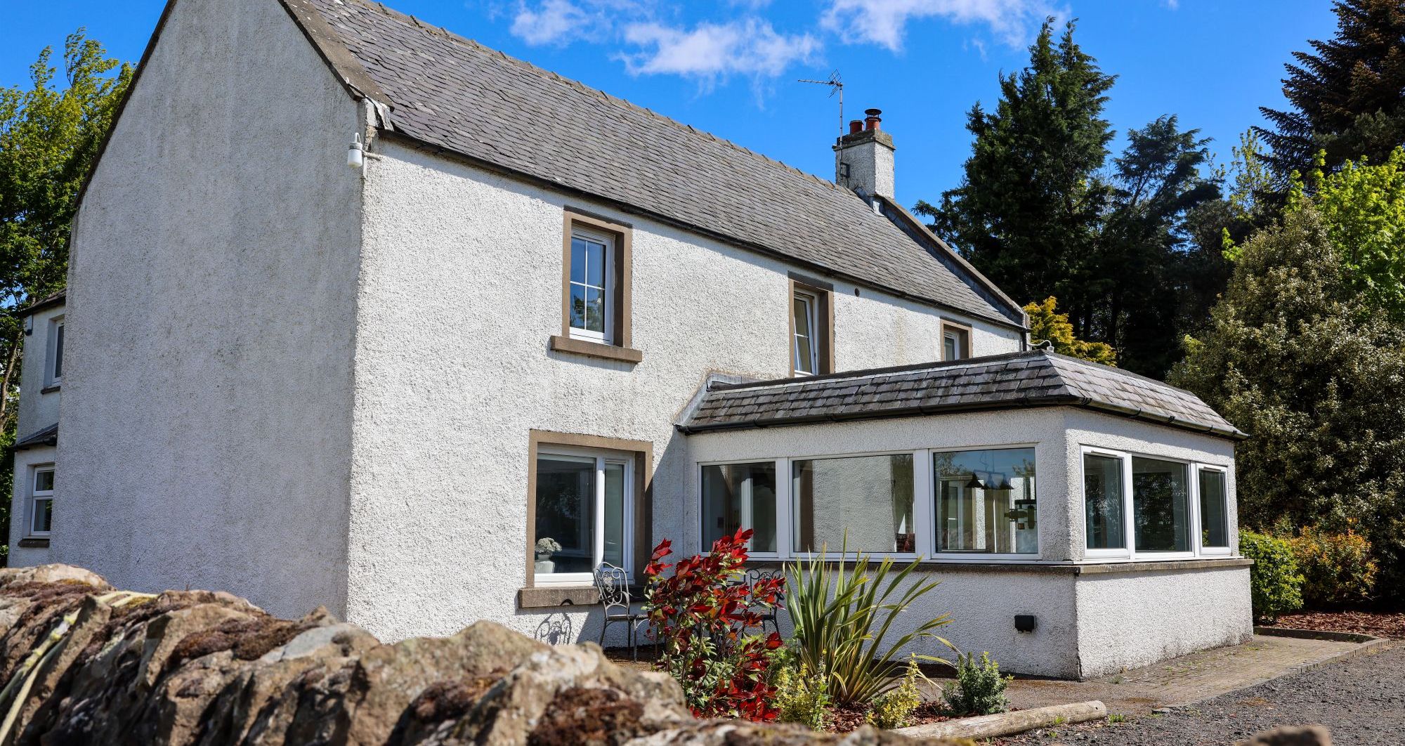 White two-story house with a grey tiled roof, a conservatory, and a stone garden wall in front.