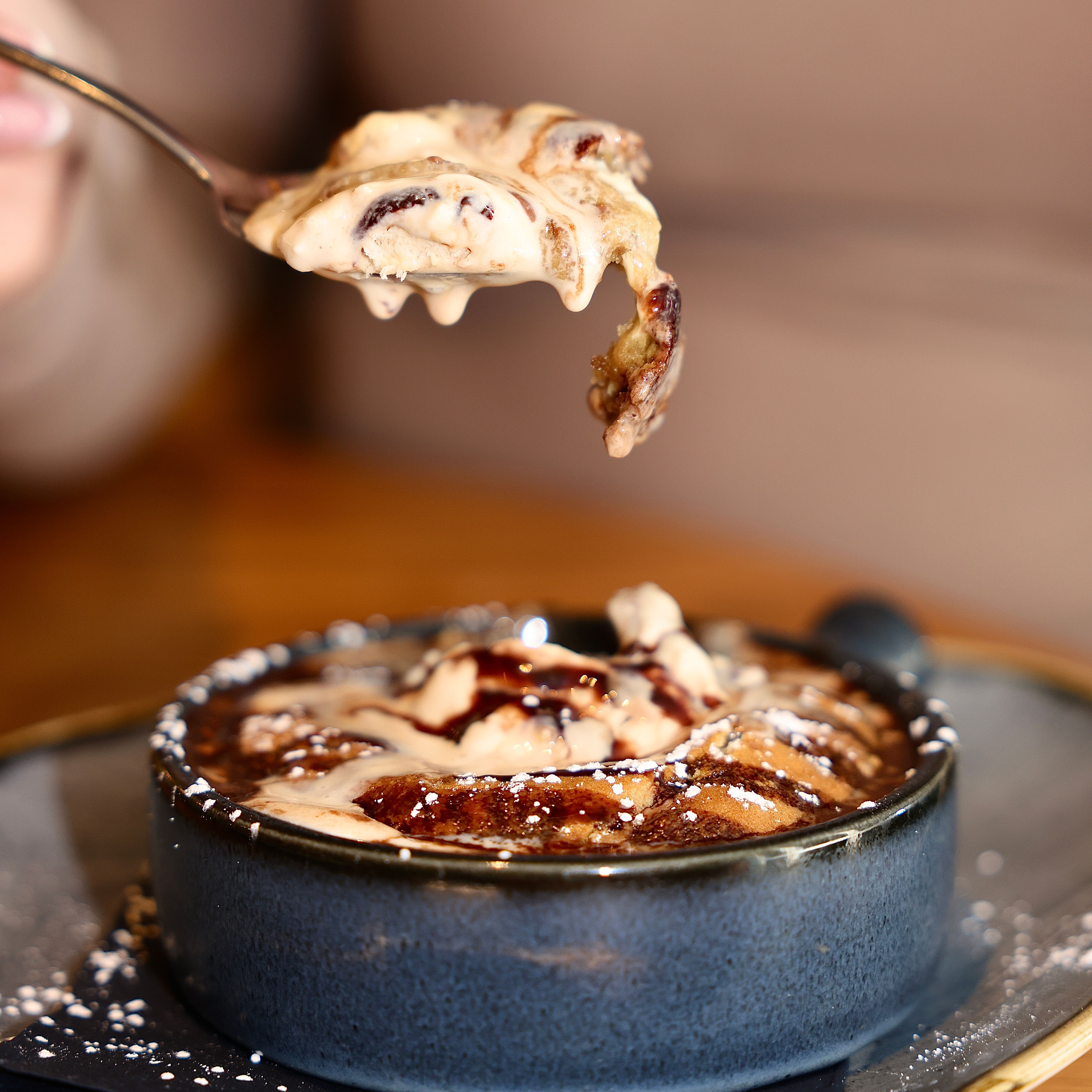 Close-up of a spoonful of dessert being lifted from a blue ceramic bowl, topped with cream and chocolate drizzle.