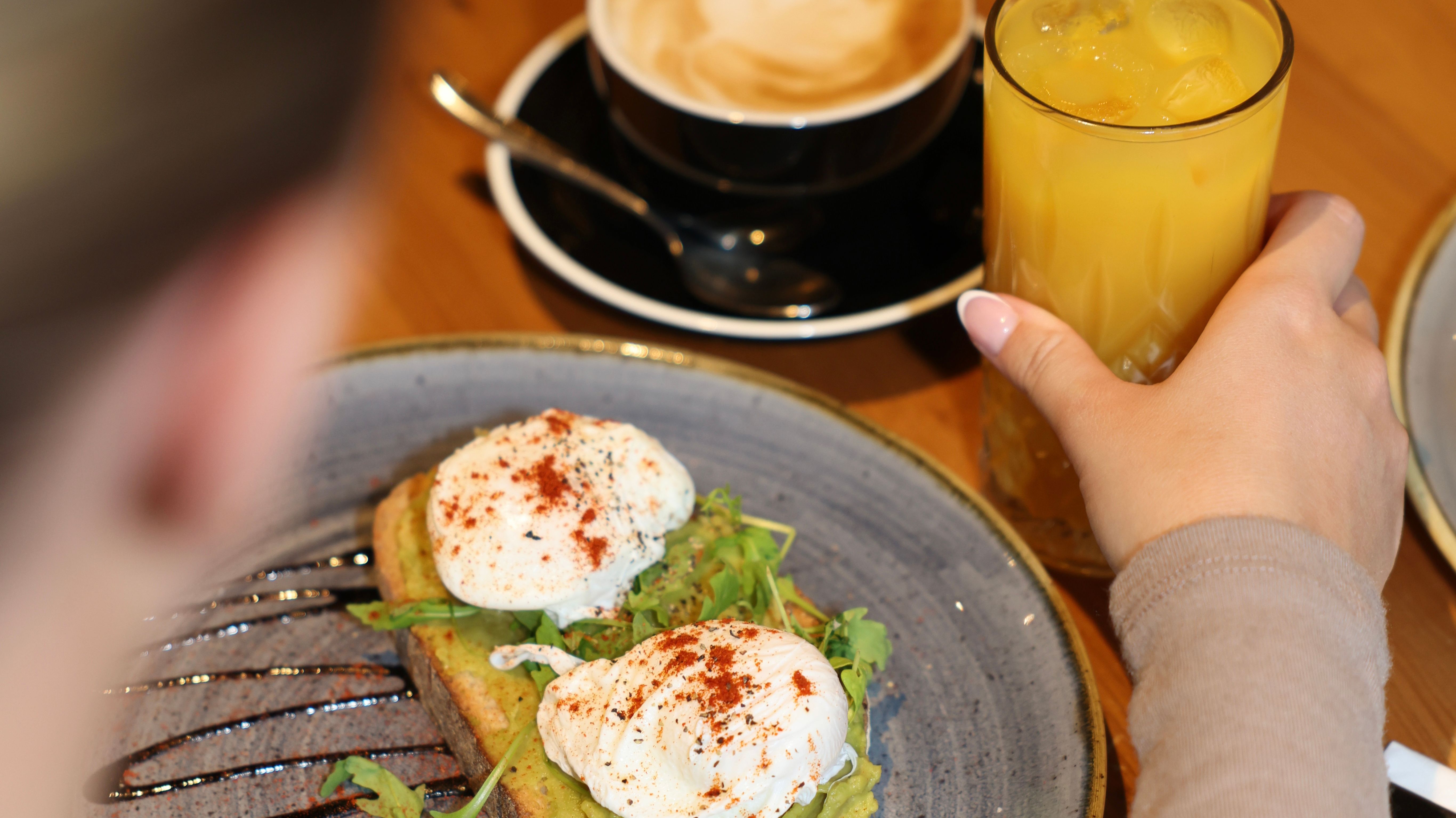 Person enjoying a breakfast of poached eggs on avocado toast, accompanied by a glass of orange juice and a cup of coffee.