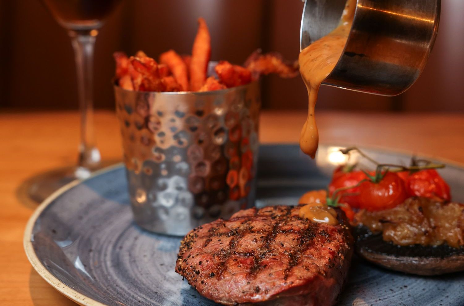 A hand pouring sauce over a grilled steak served with fries, roasted vegetables, and a cocktail.