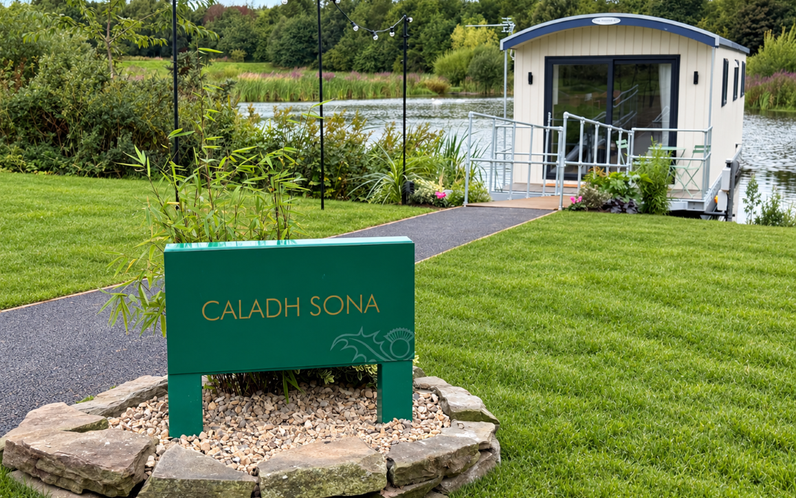 Modern floating cabin named Caladh Sona on a lake with a green sign in the foreground and landscaped garden surroundings.