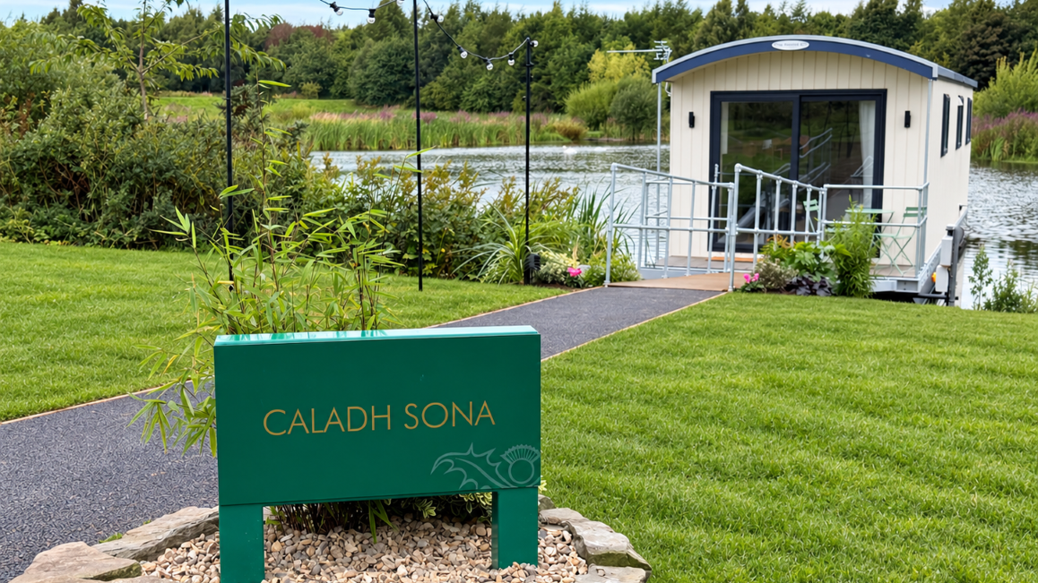 Modern floating cabin named Caladh Sona on a lake with a green sign in the foreground and landscaped garden surroundings.