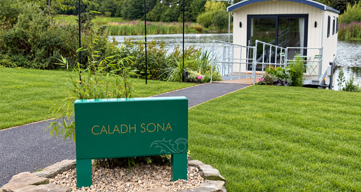 Modern floating cabin named Caladh Sona on a lake with a green sign in the foreground and landscaped garden surroundings.
