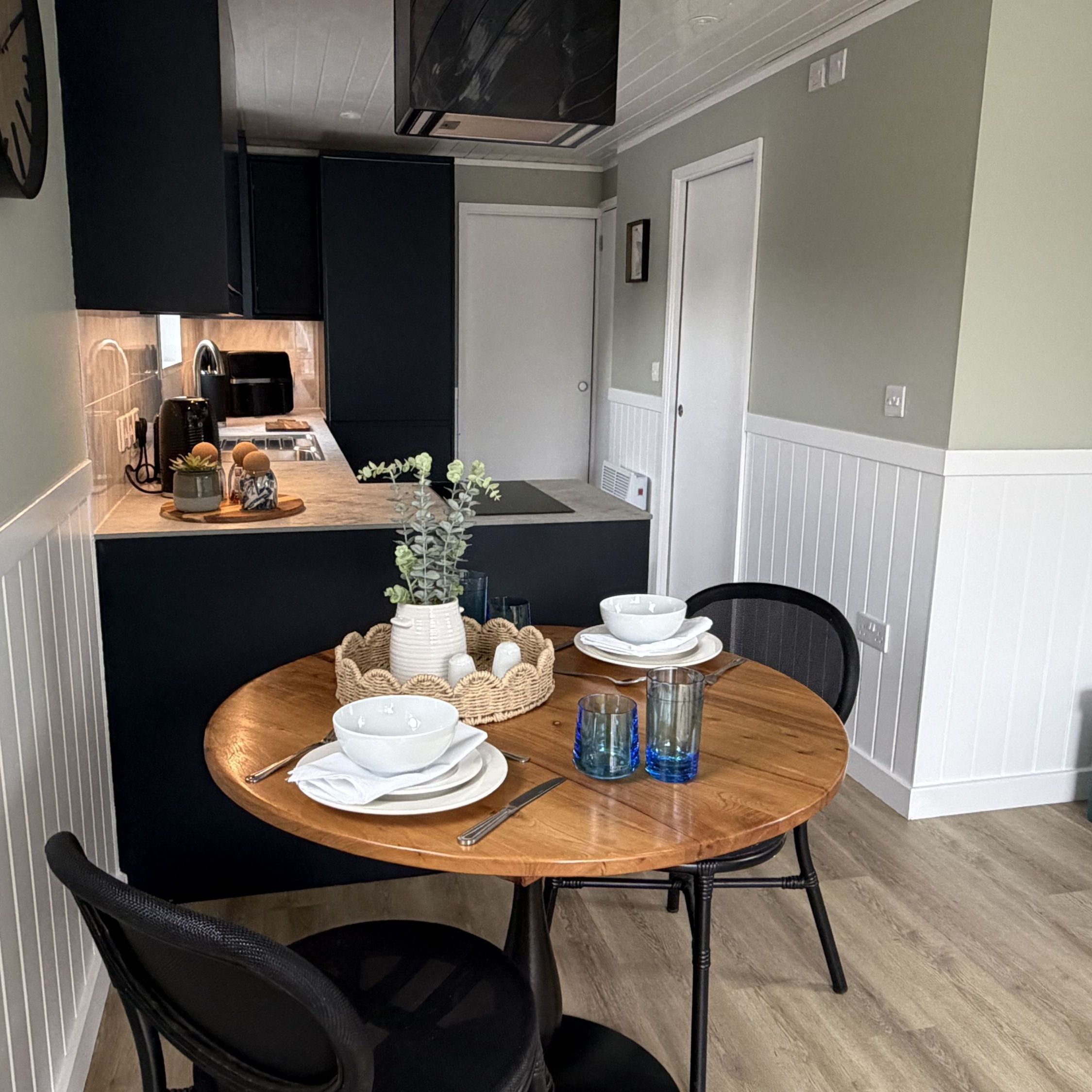 Modern kitchen and dining area with a round wooden table set for two people, dark cabinets, and light green walls with white wainscoting.