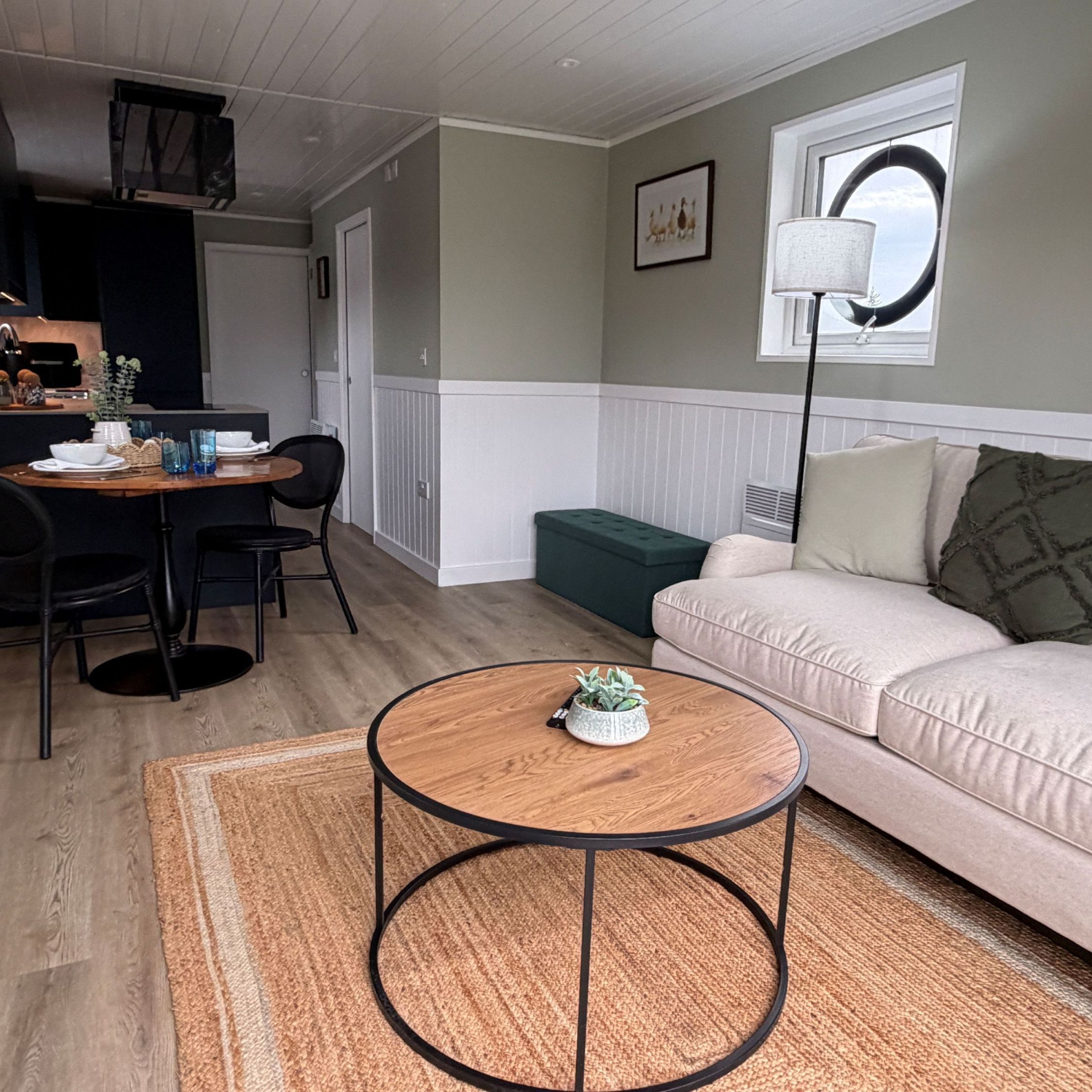 Modern open-plan living and kitchen area with a beige sofa, round coffee table, dining table for two, and compact kitchen.
