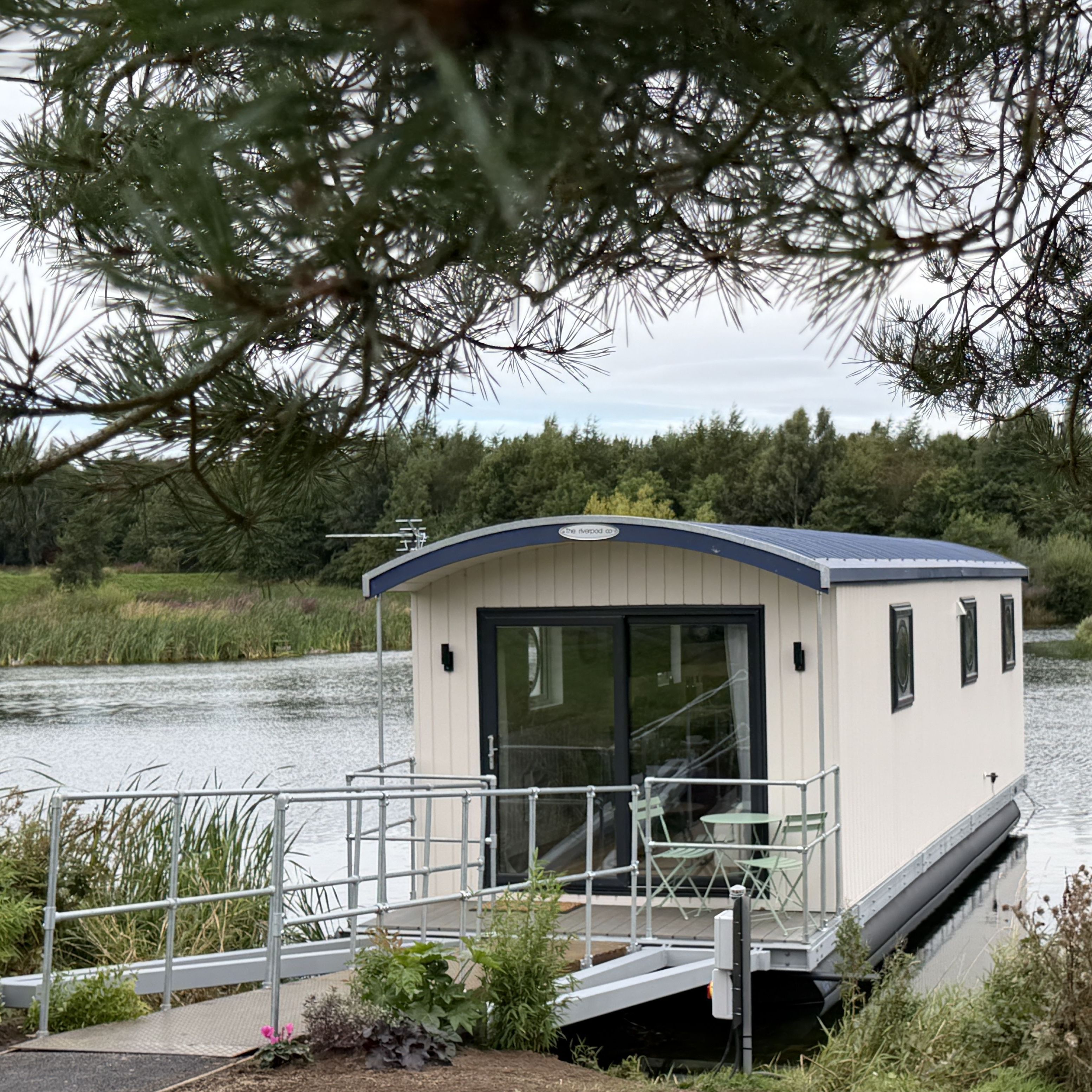 Floating house with a patio on a calm lake surrounded by greenery and trees.