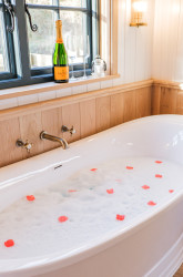 Bath with flower petals and bottle of champagne on windowsill