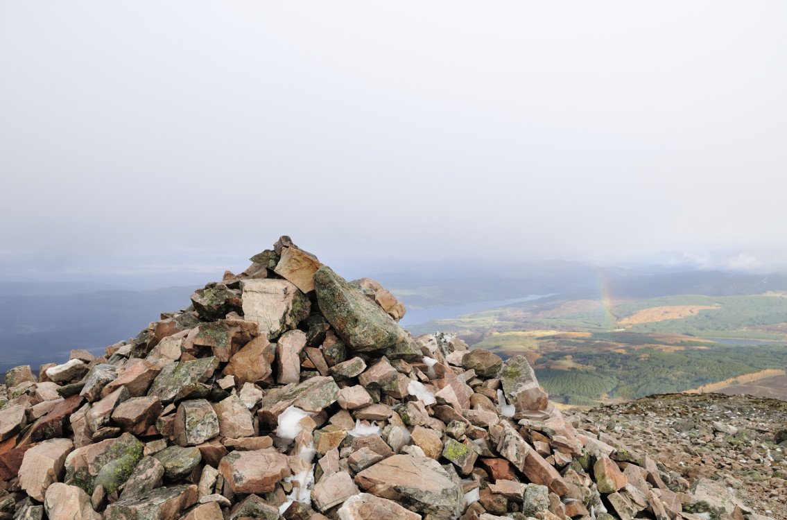 Rock cairn atop a munro with a distant view of a rainbow over the landscape.