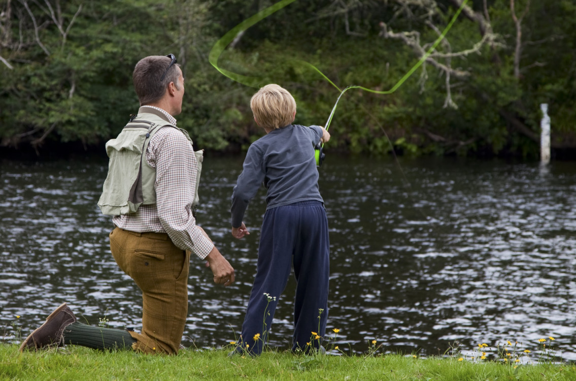 Father and son fly fishing together by a riverbank surrounded by greenery.