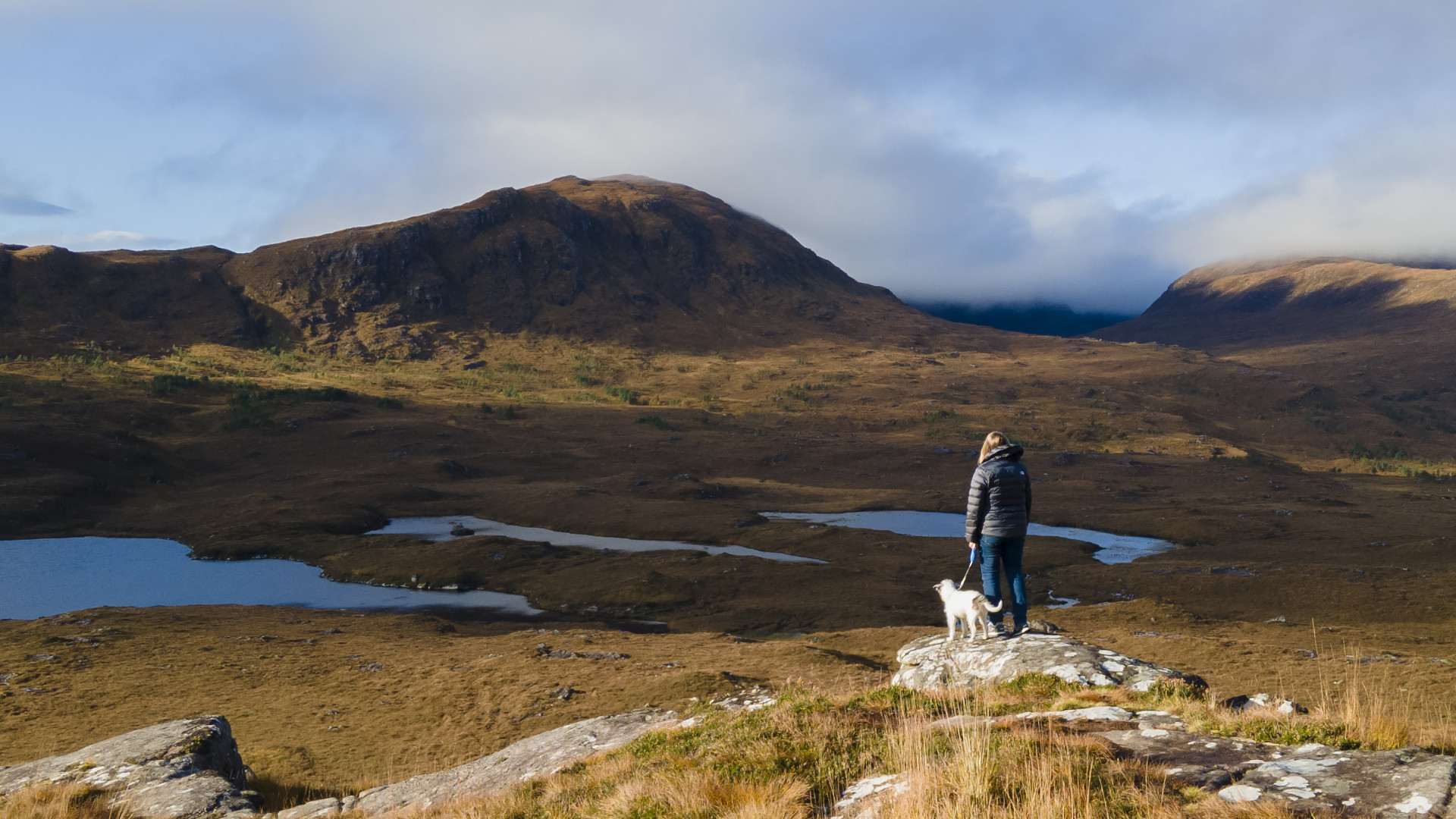 Hiker with a dog standing on a rocky hill, overlooking a scenic mountain landscape.